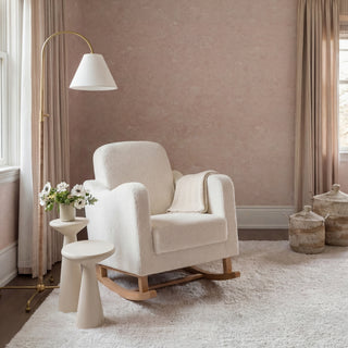 White rocking chair with a soft throw blanket, beside a small table with flowers, in a light-colored room.